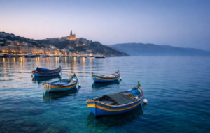 Gozo Travel Guide: The Best Things to Do, Gozo Sightseeing & Places to Visit Mgarr Harbour in Gozo at blue hour with traditional Maltese fishing boats floating on clear turquoise water and the illuminated church silhouette on the hillside.