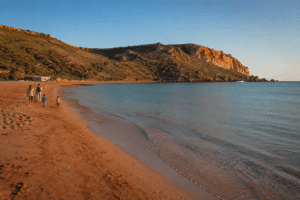 Best Beaches for Families in Gozo Families walking along the red sand shoreline of Ramla Bay in Gozo during calm early morning Mediterranean light.